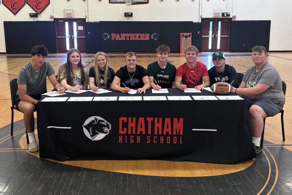 six boys and two girls singing letters while seated at table draped with Chatham High School logo